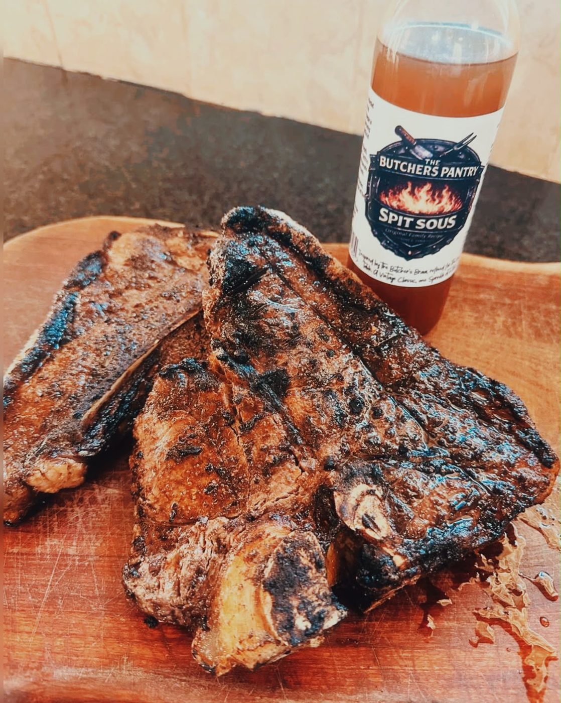 Grilled beef chops on a wooden board beside a bottle of Spit Sous